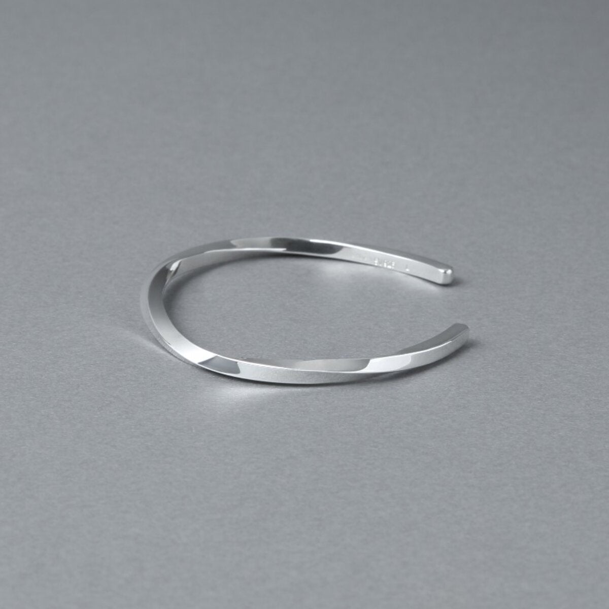 シルバー925バングル「21-B-005」(幅3mm)を、開口部を右側に向けてグレー背景で斜め上から撮影。ねじりのあるフォルムと鏡面仕上げ、内側の刻印が見える商品写真。周囲に余白を残した構図。