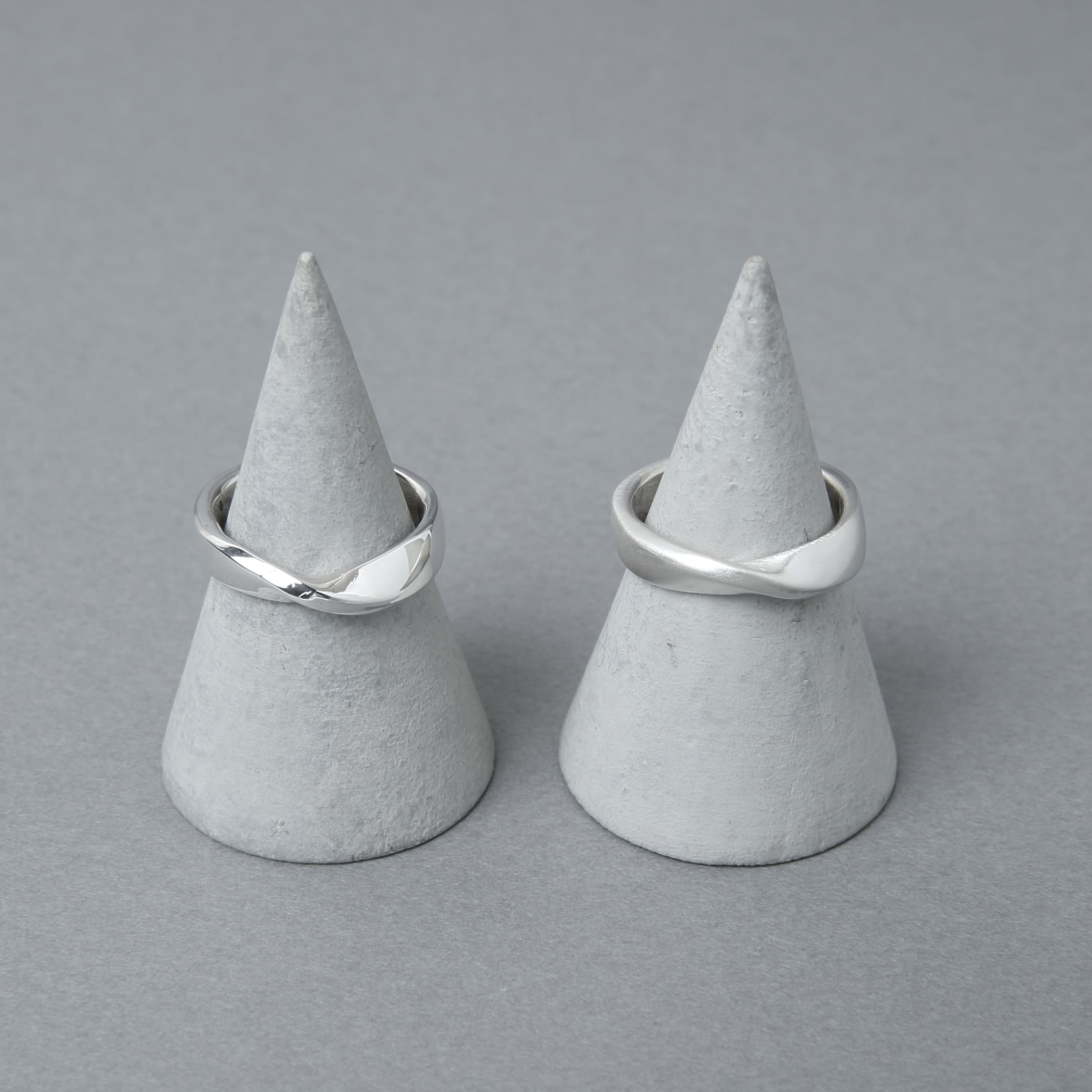 2つのシルバー925製リングがグレーのコーン型ディスプレイにセットされている。リングはシンプルなデザインで、控えめなツイストが加えられ、背景もシンプルなグレーで統一されている。