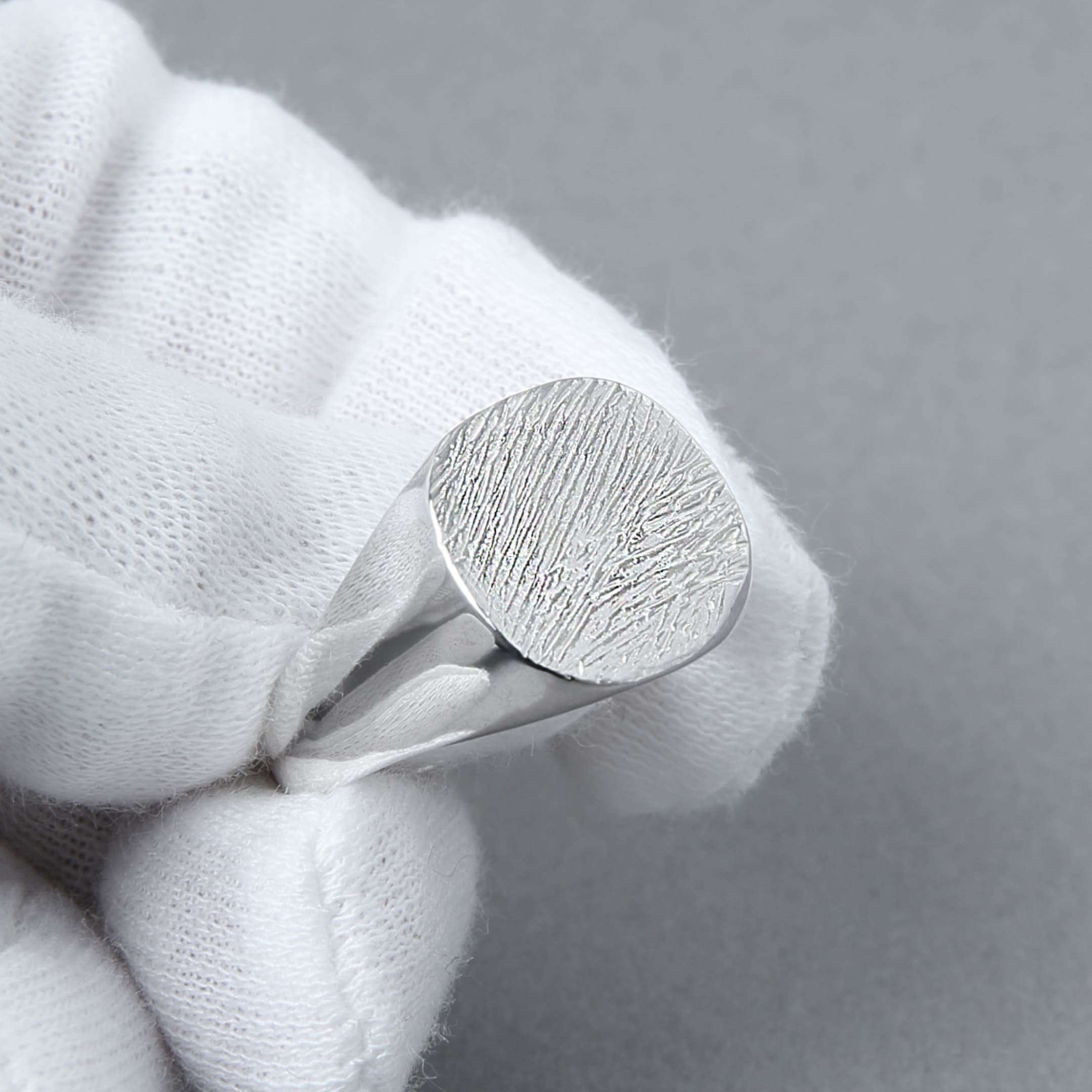 白い手袋をはめた手が、木目模様のようなテクスチャが施されたシルバー925のラウンドシグネットリングを持っている。上品な質感と滑らかな鏡面仕上げが際立つ。