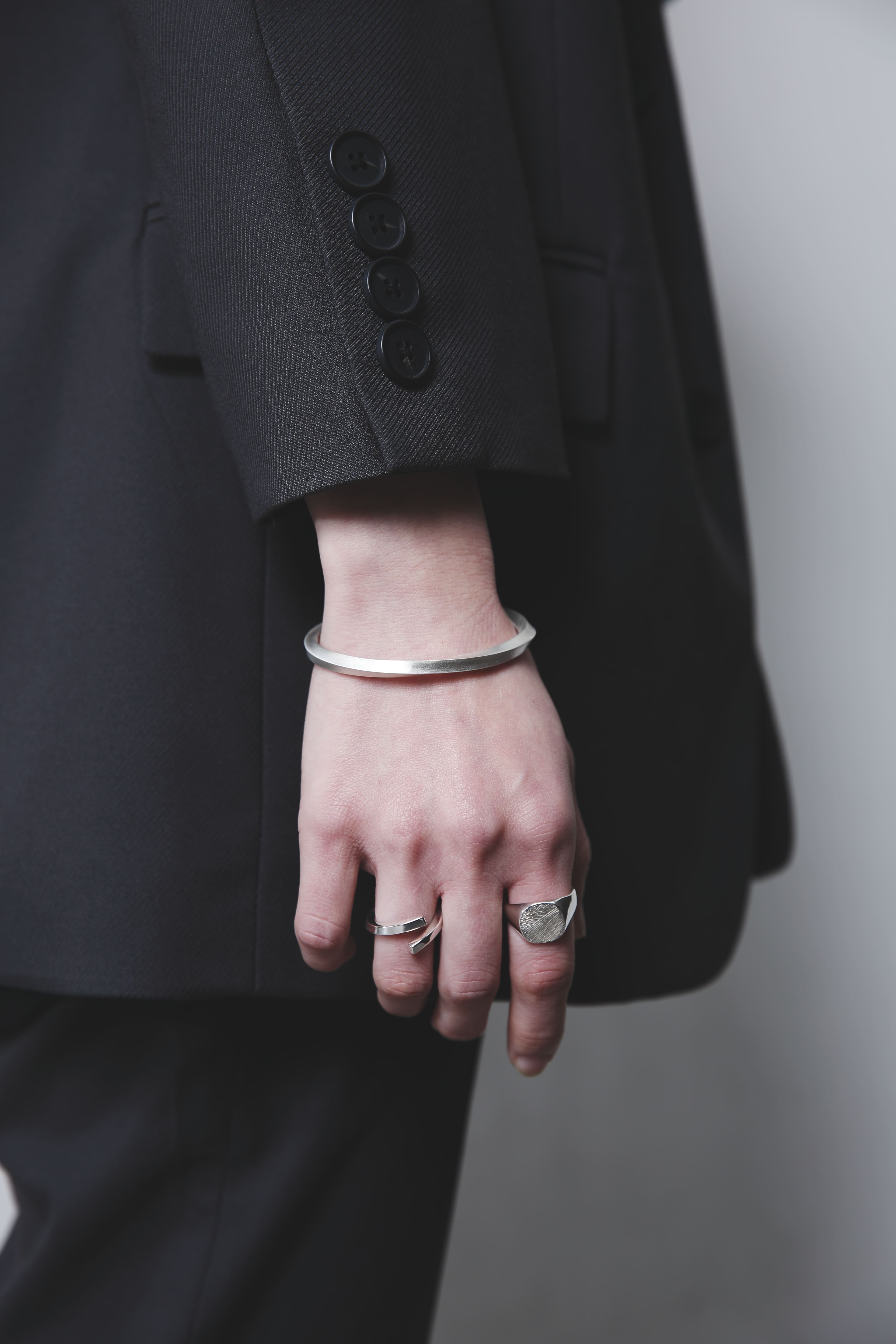 黒のジャケットを着用した人物の手元。木目模様のシルバーリングとオープンリング、そしてマットな質感のシルバーバングルを重ね付けしている。シンプルな装いに洗練された輝きが映える。