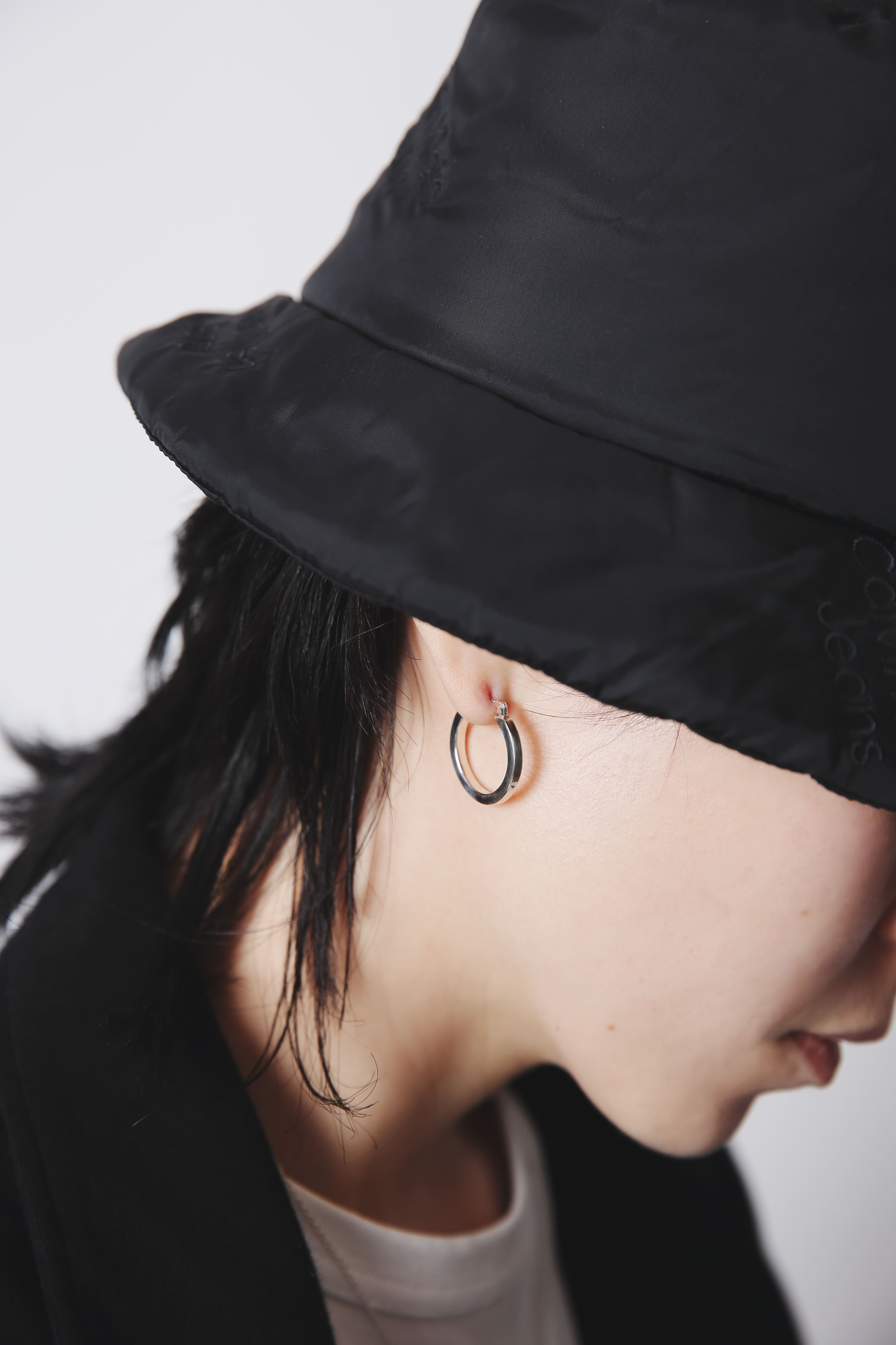 黒いバケットハットのつばの下から見える耳に、小ぶりなシルバーフープピアスが装着されているクローズアップカット。