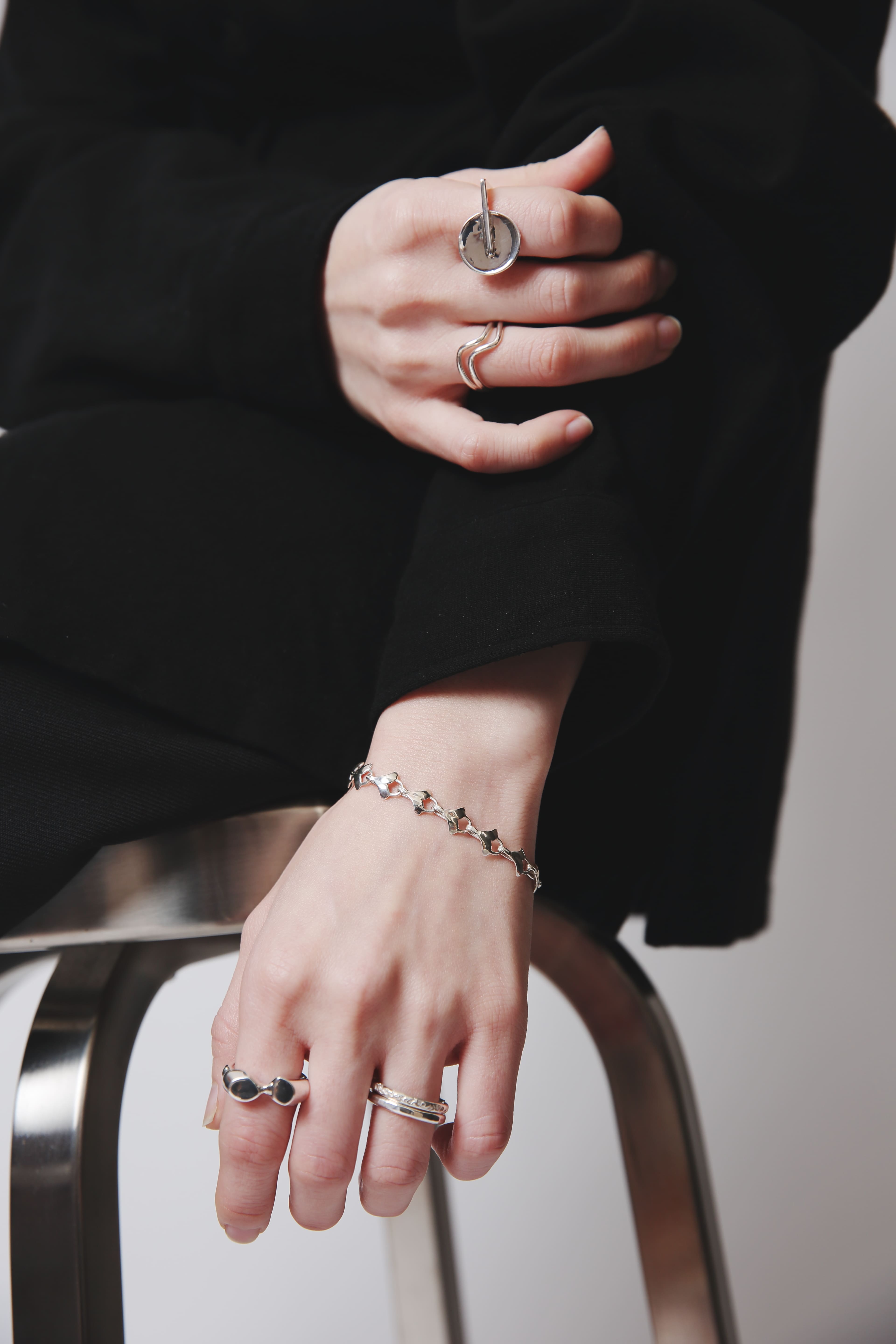 椅子に肘を置き、手を組むように添えた姿から覗く、光沢のあるイチョウの葉モチーフブレスレット。洗練されたモノトーンスタイルの中で印象的に輝く。
