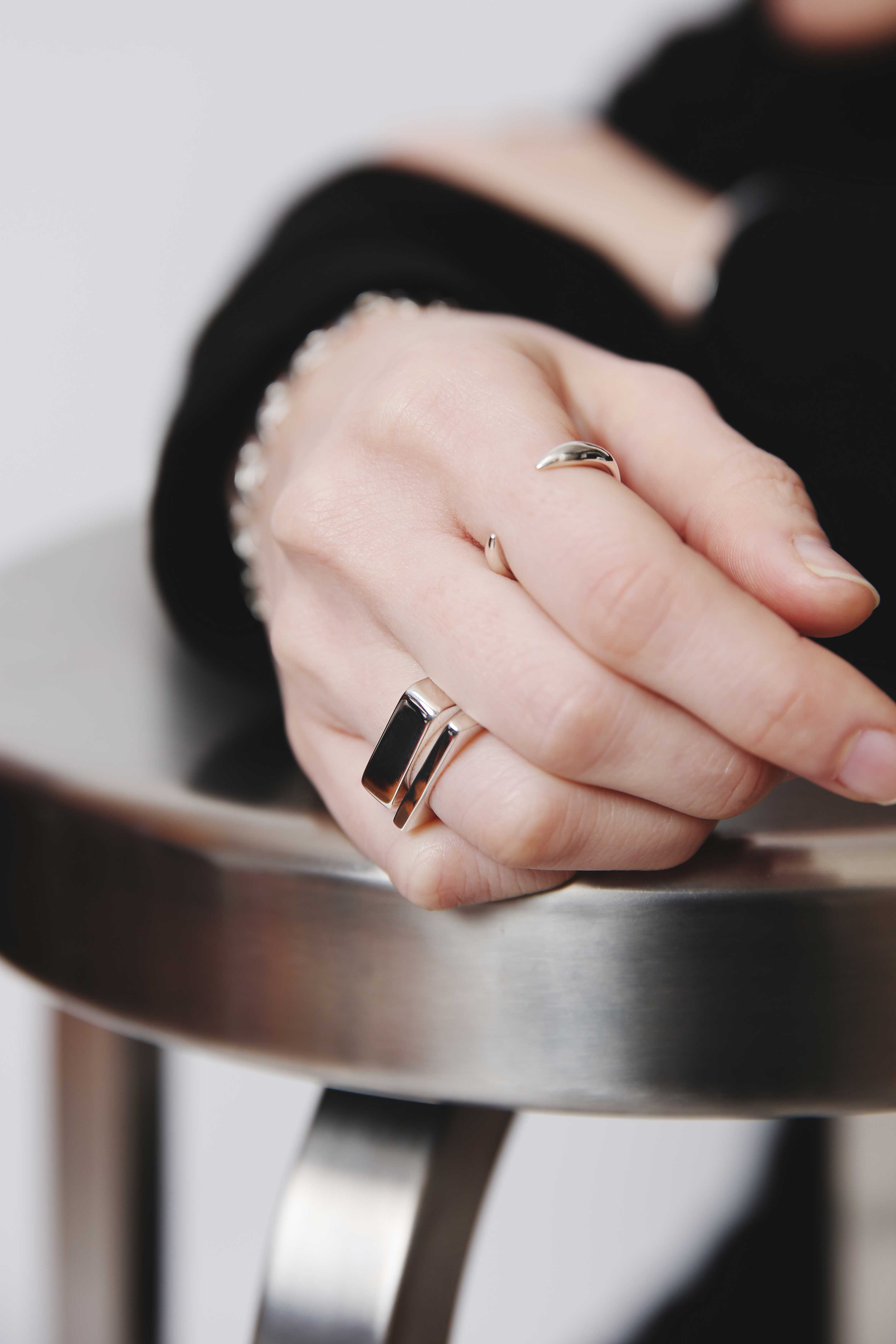 スツールにもたれる手元に輝くNine Nineのシルバー925製スクエアリング2連とミニマルオープンリング—艶感際立つブラックコーデ