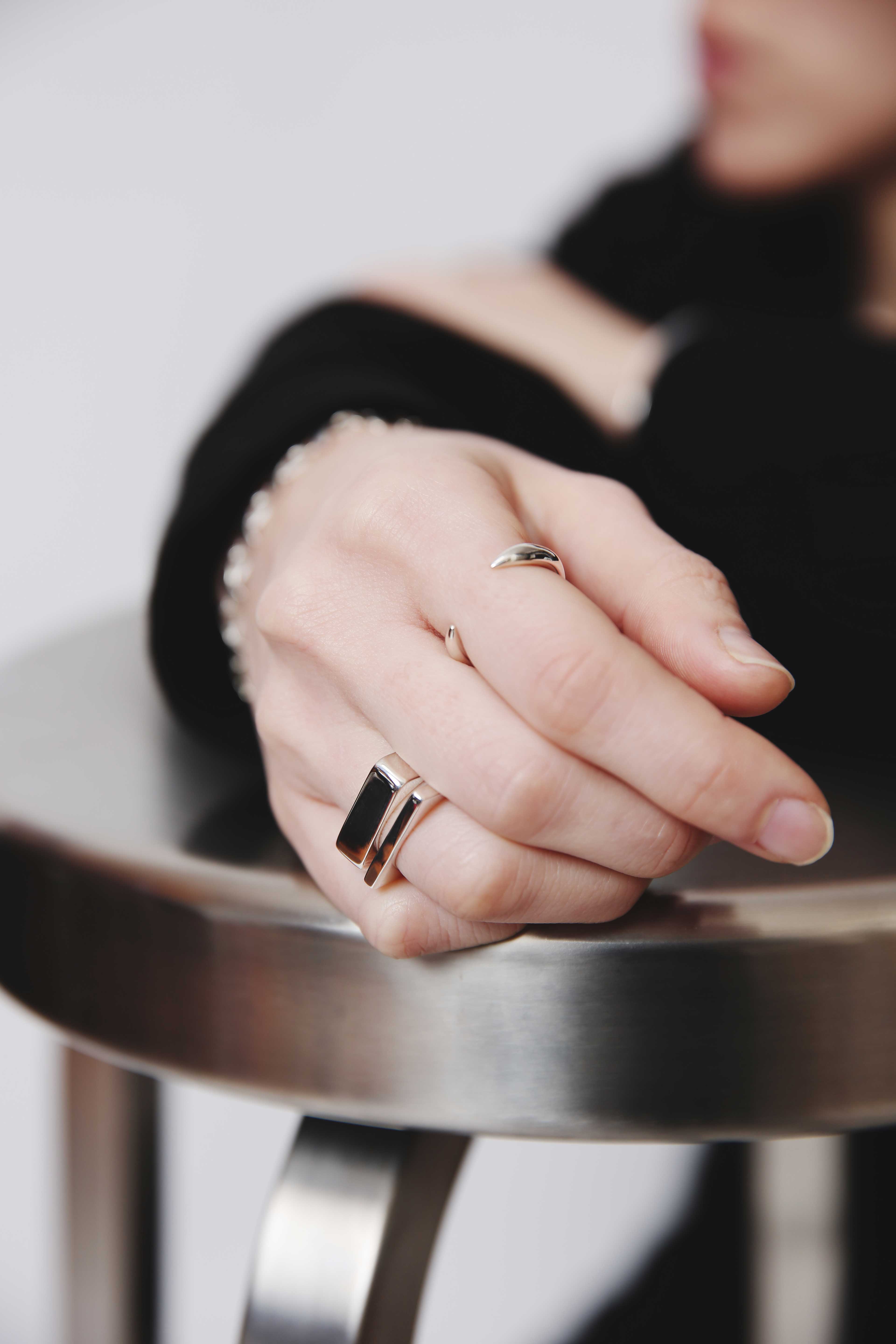 2本のシルバーリングを着用した指先をフォーカスしたイメージ。ブラックトップスとシンプルなメタルチェアを背景にしたスタイリッシュな構図。