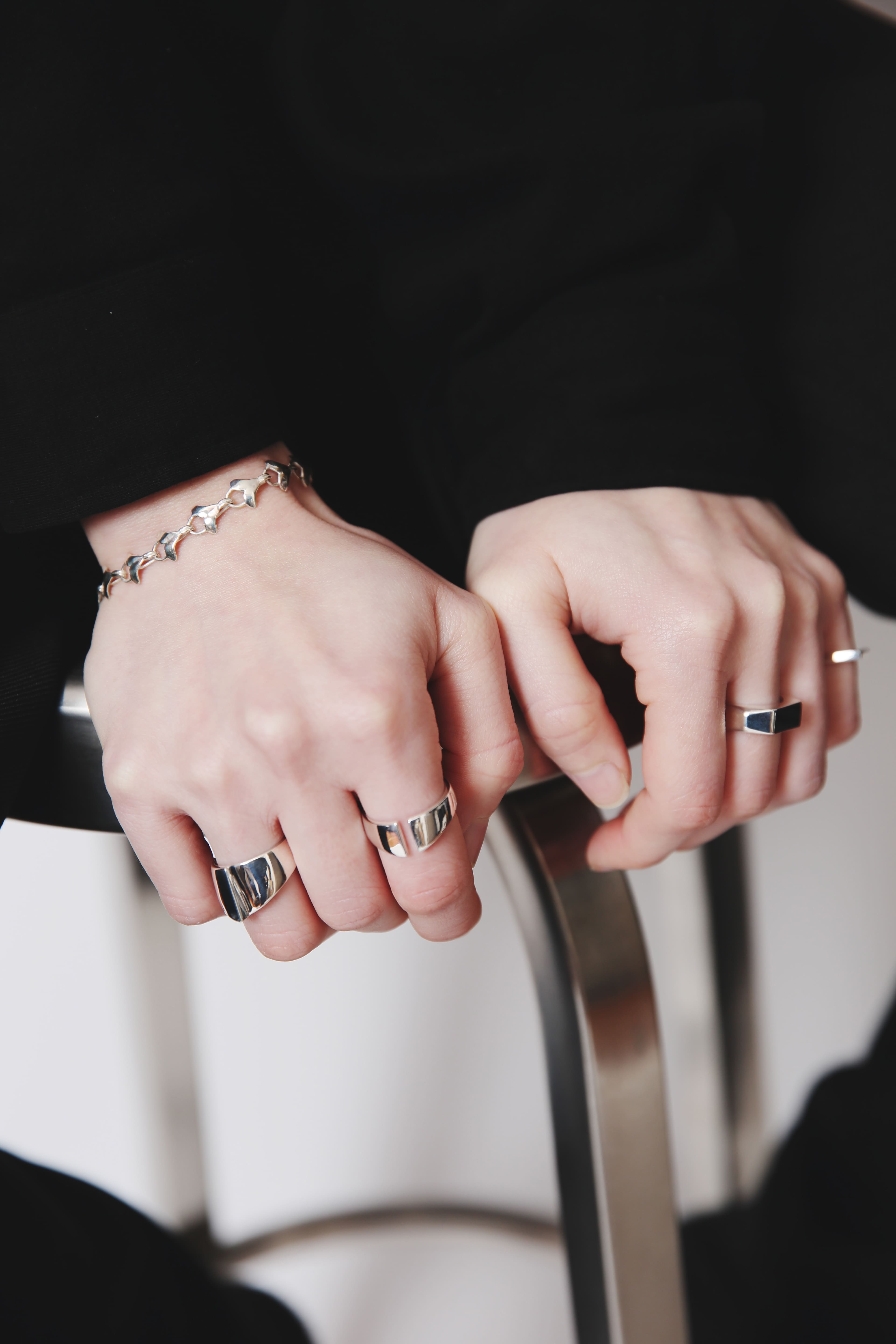 椅子の金属フレームに手を置いた両手のアップ。右手首にイチョウの葉モチーフのシルバーブレスレット、左手の人差し指と中指にV字の切り込みが入った幅広リング、左手人差し指にプレートリングを着用