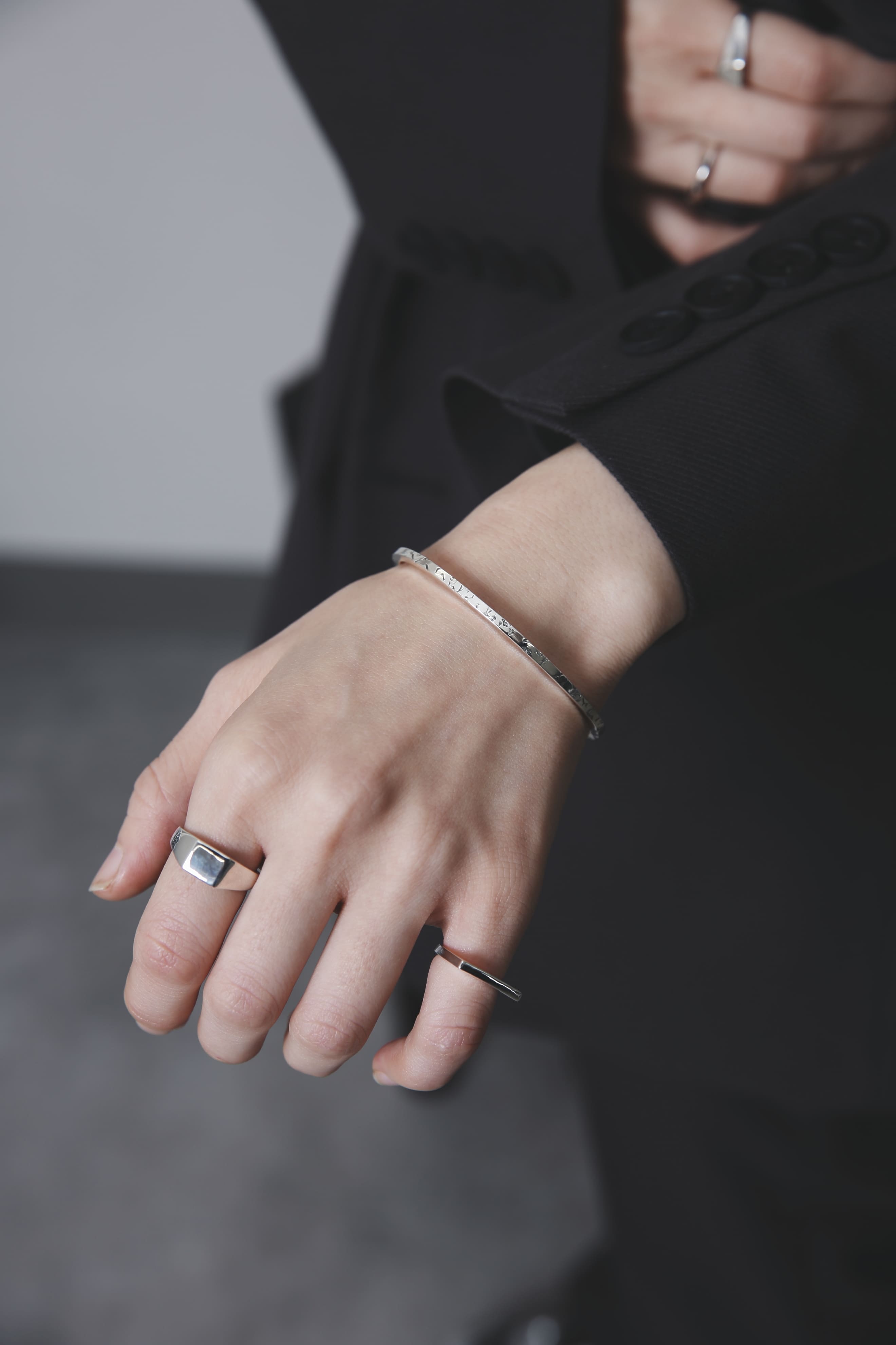 手の甲をやや前に出した構図で、バングルと2本のリングが光を受けて輝く。クラシックな黒の装いに、無駄のないシルバージュエリーが映えるカット。