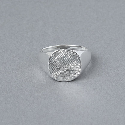 グレーの背景上に置かれた、丸いトップに繊細な木目模様が刻まれたシルバー925リング。立体的な陰影が素材の輝きを引き立てている。