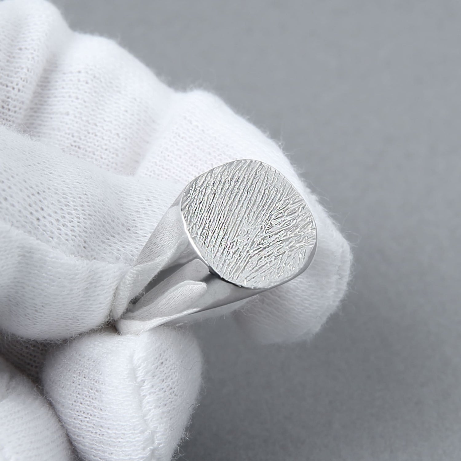 白い手袋をはめた手が、木目模様のようなテクスチャが施されたシルバー925のラウンドシグネットリングを持っている。上品な質感と滑らかな鏡面仕上げが際立つ。