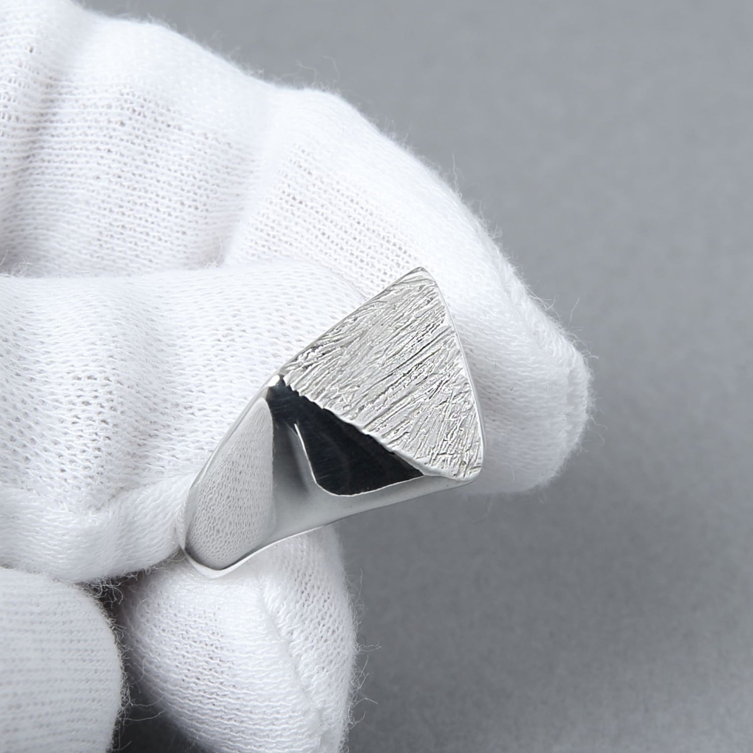 白い手袋を着けた手が、木目模様の三角形シグネットリングを持つ様子。職人による繊細なテクスチャと輝きが際立つディテールショット。