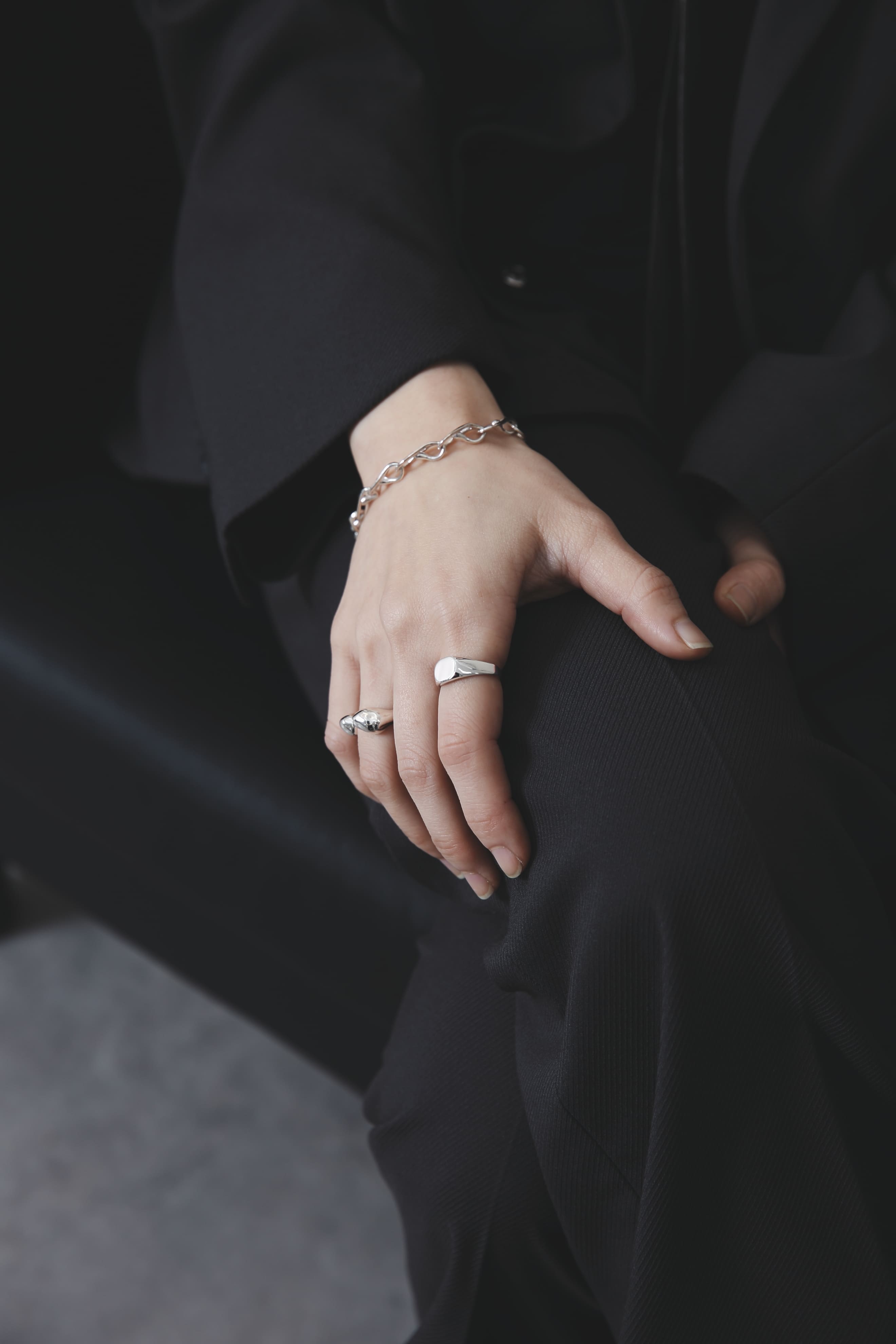 黒いジャケットを着た人物の手元。丸型トップのシグネットリングと、立体的なデザインのリング、チェーンブレスレットを重ね付け。ミニマルな装いに素材の輝きが際立つ。