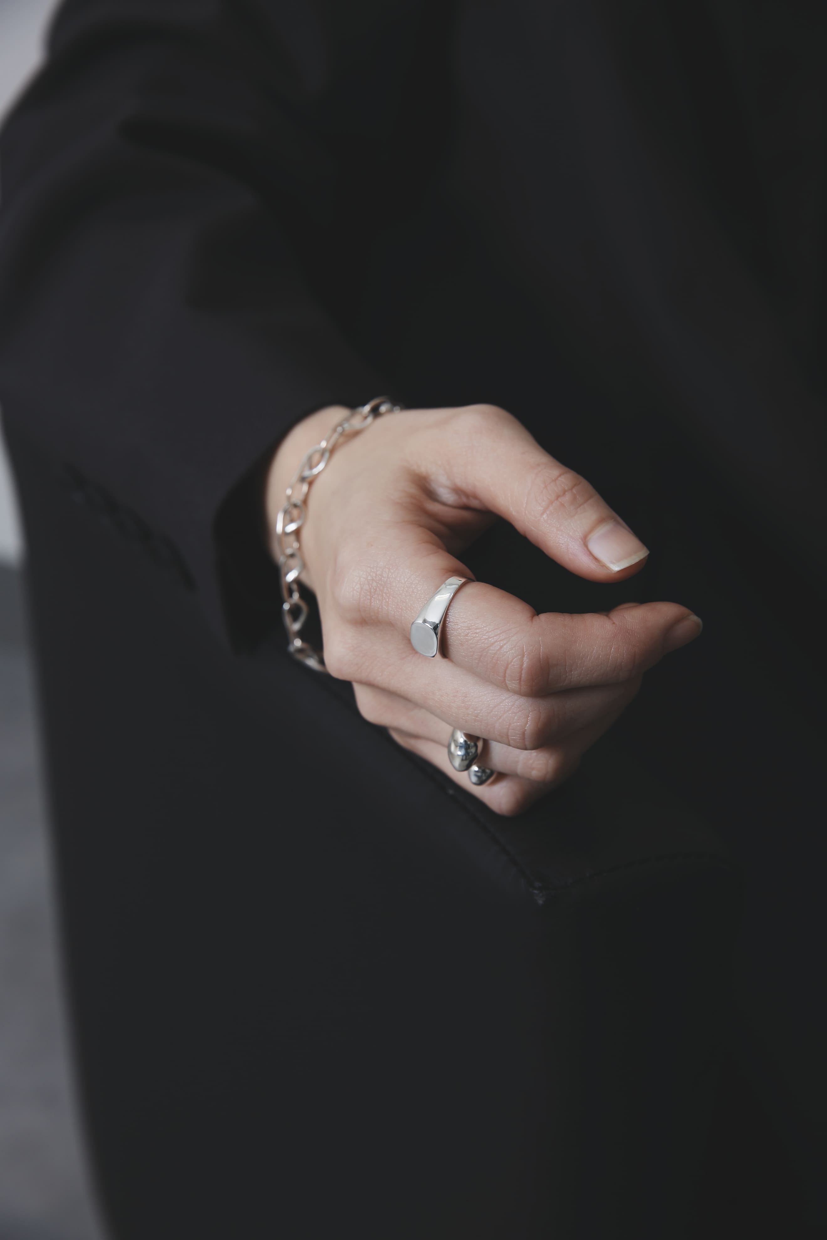 黒い椅子の背に手を預けた姿。丸型シシグネットリングの鏡面仕上げが光を柔らかく反射し、チェーンブレスレットとのバランスが美しい。