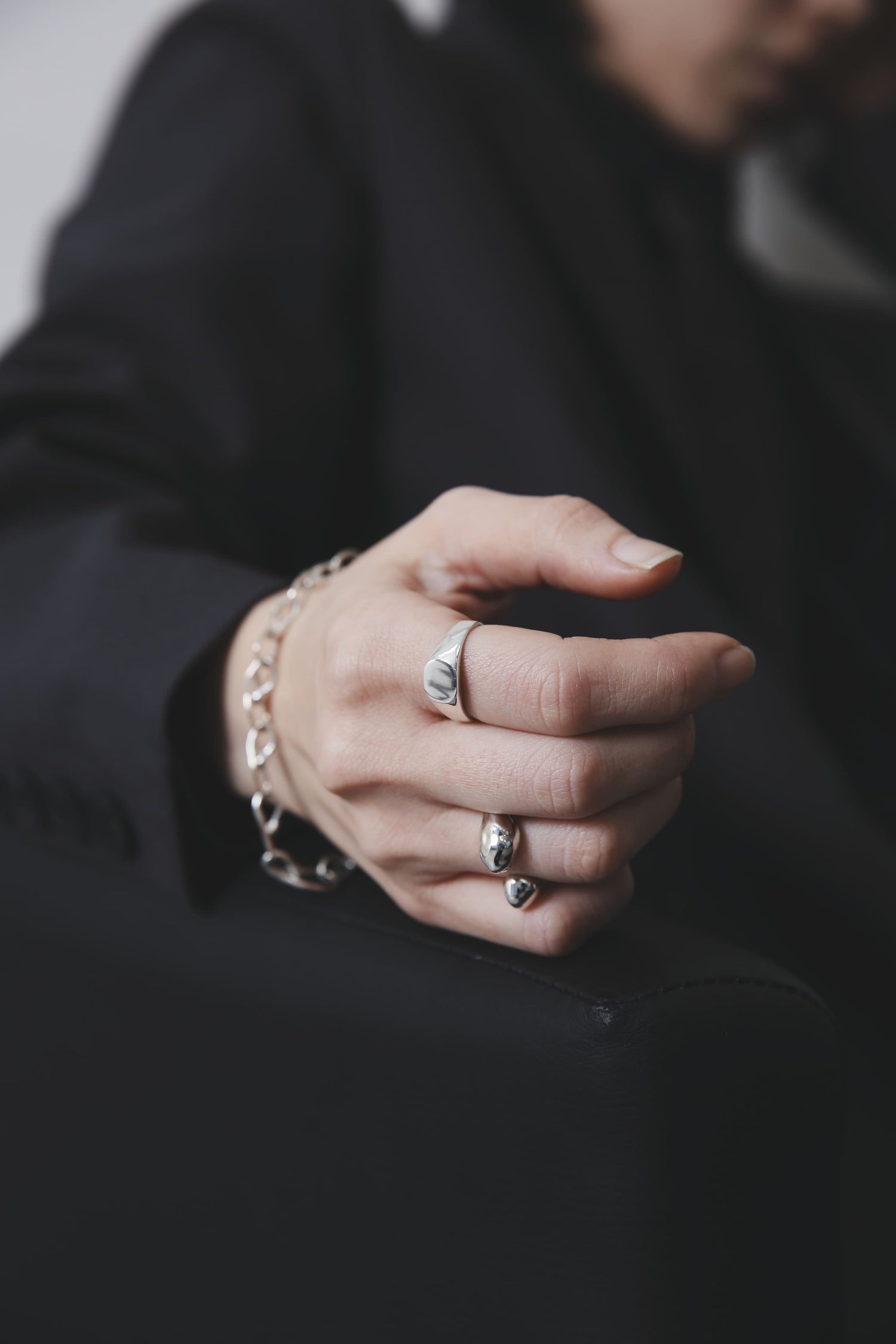 ソファの縁に手を置き、軽く拳を握ったポーズ。ミニマルなシグネットリングとドロップ状モチーフのリングが、造形的な美しさを強調している