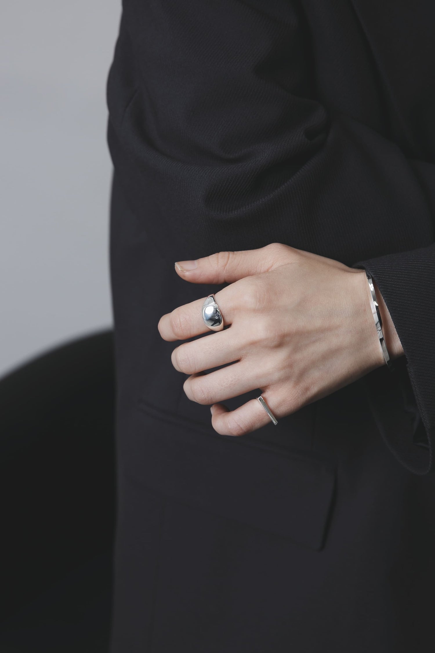 黒いジャケットの袖口を軽く握る手元のアップ。鏡面仕上げの丸型シルバーリングと、シャープなバングルが指先を洗練された印象に見せている