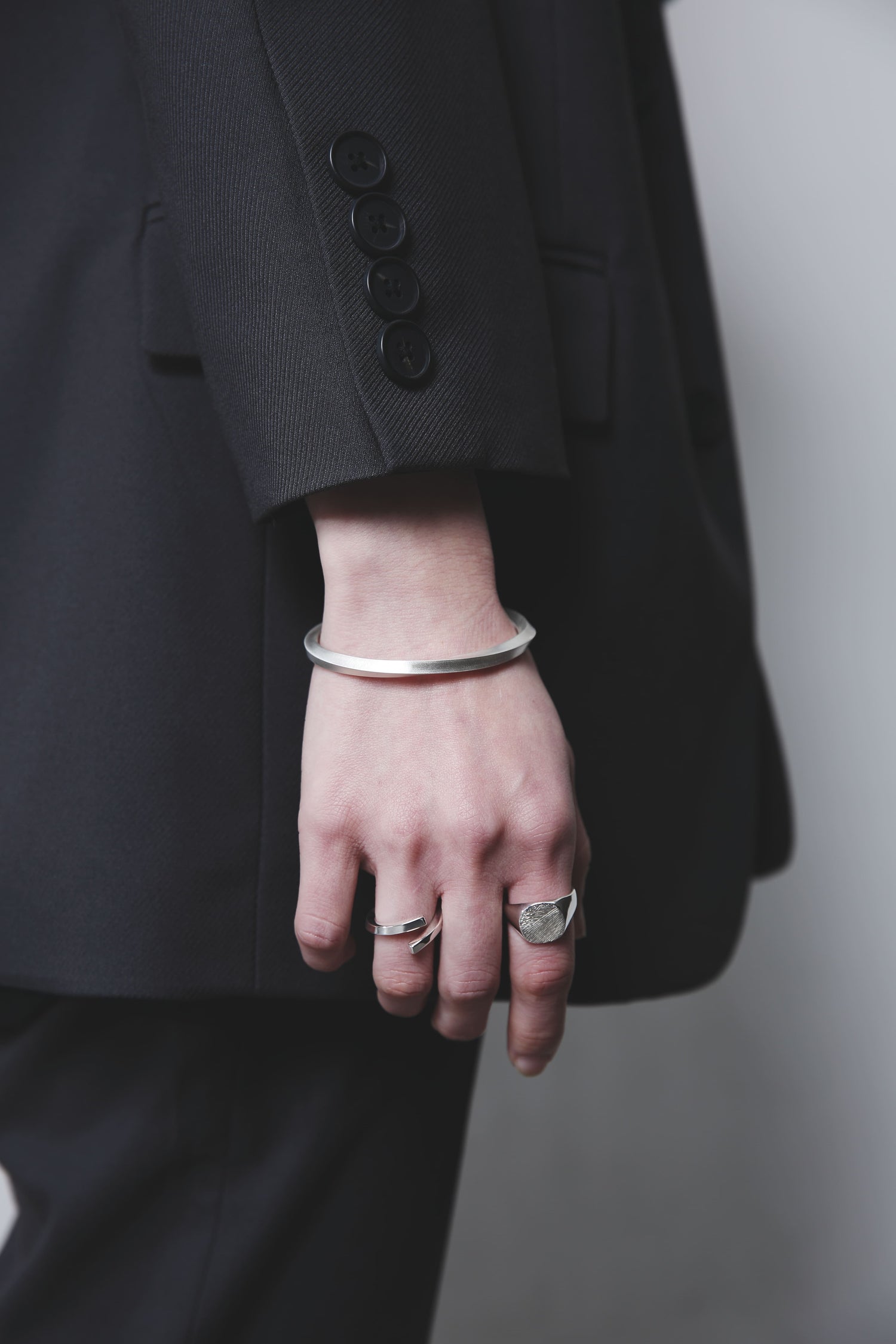 黒のジャケットを着用した人物の手元。木目模様のシルバーリングとオープンリング、そしてマットな質感のシルバーバングルを重ね付けしている。シンプルな装いに洗練された輝きが映える。
