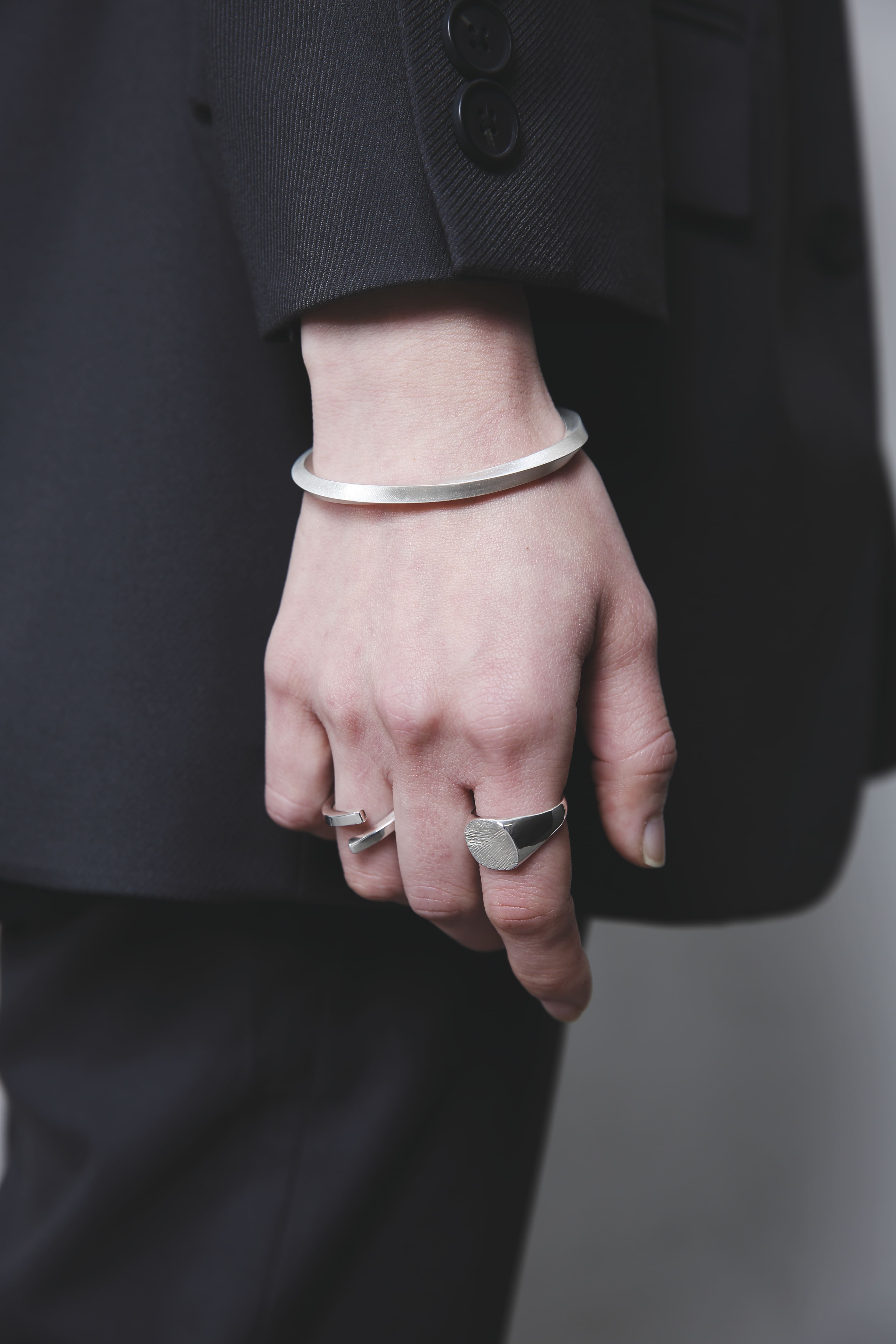 黒いジャケットの袖口から覗くシルバーバングルと、木目模様のシルバーリング。直線的なフォルムと滑らかな光沢がスタイリッシュな印象を与える。