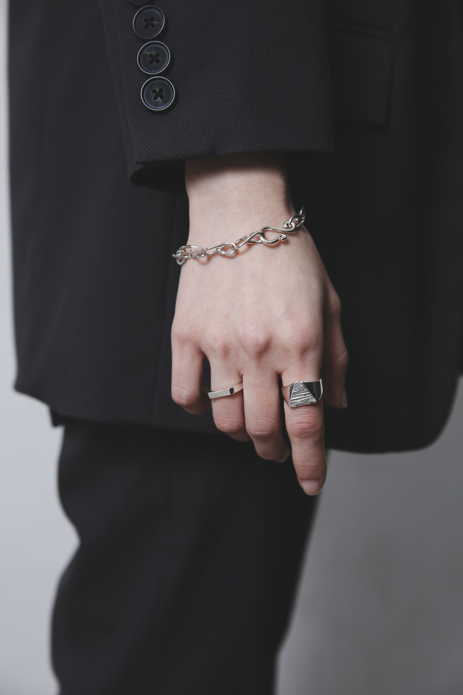 手首のチェーンブレスレットと三角トップのシグネットリングを中心に撮影。シルバーの質感と自然光の反射が美しく調和している。