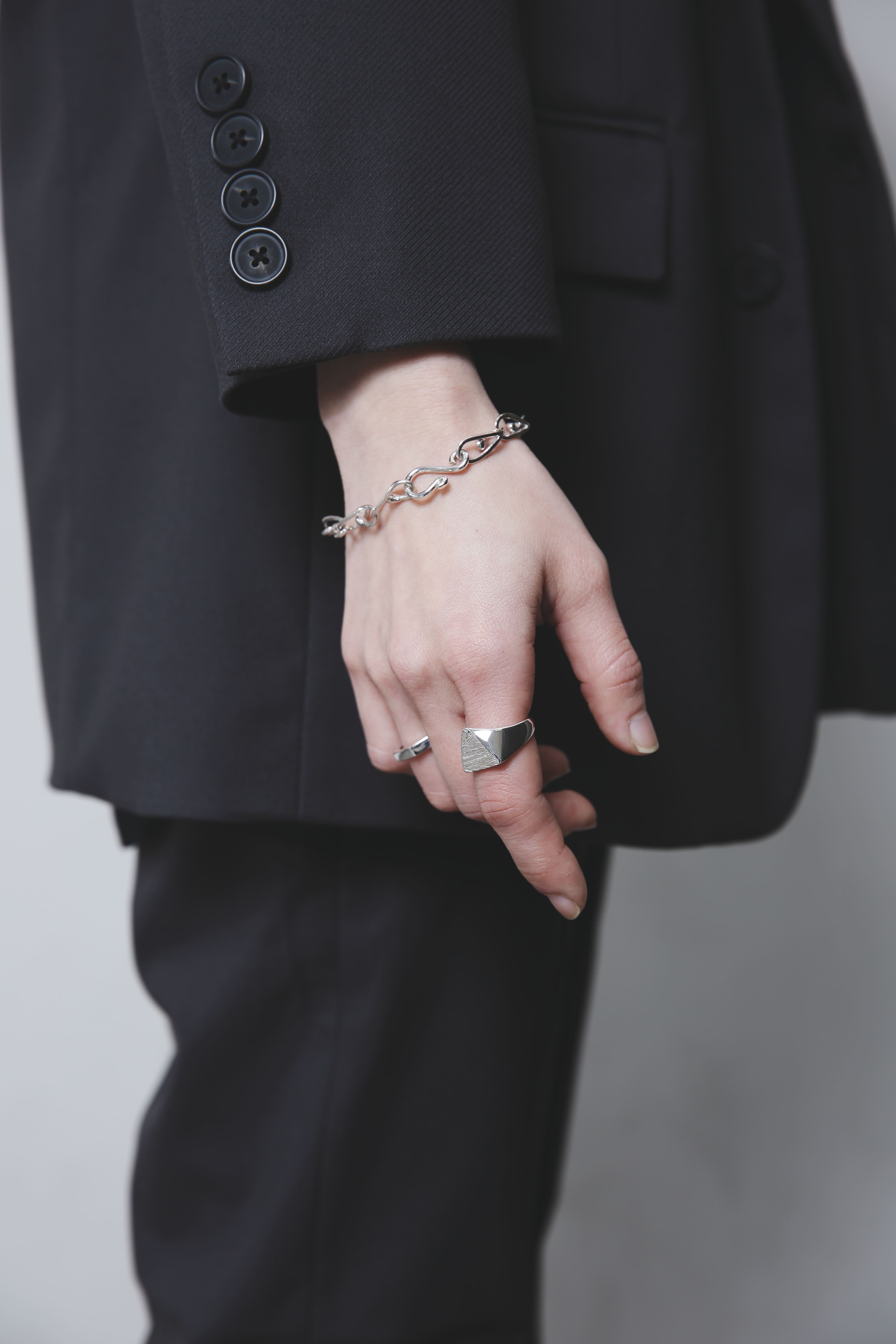 黒いスーツの袖口から覗く手元。滑らかな曲線を描くチェーンブレスレットと、幾何学的なシルバーリングのコントラストが印象的。