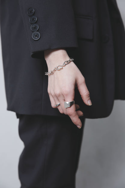 黒いスーツの袖口から覗く手元。滑らかな曲線を描くチェーンブレスレットと、幾何学的なシルバーリングのコントラストが印象的。