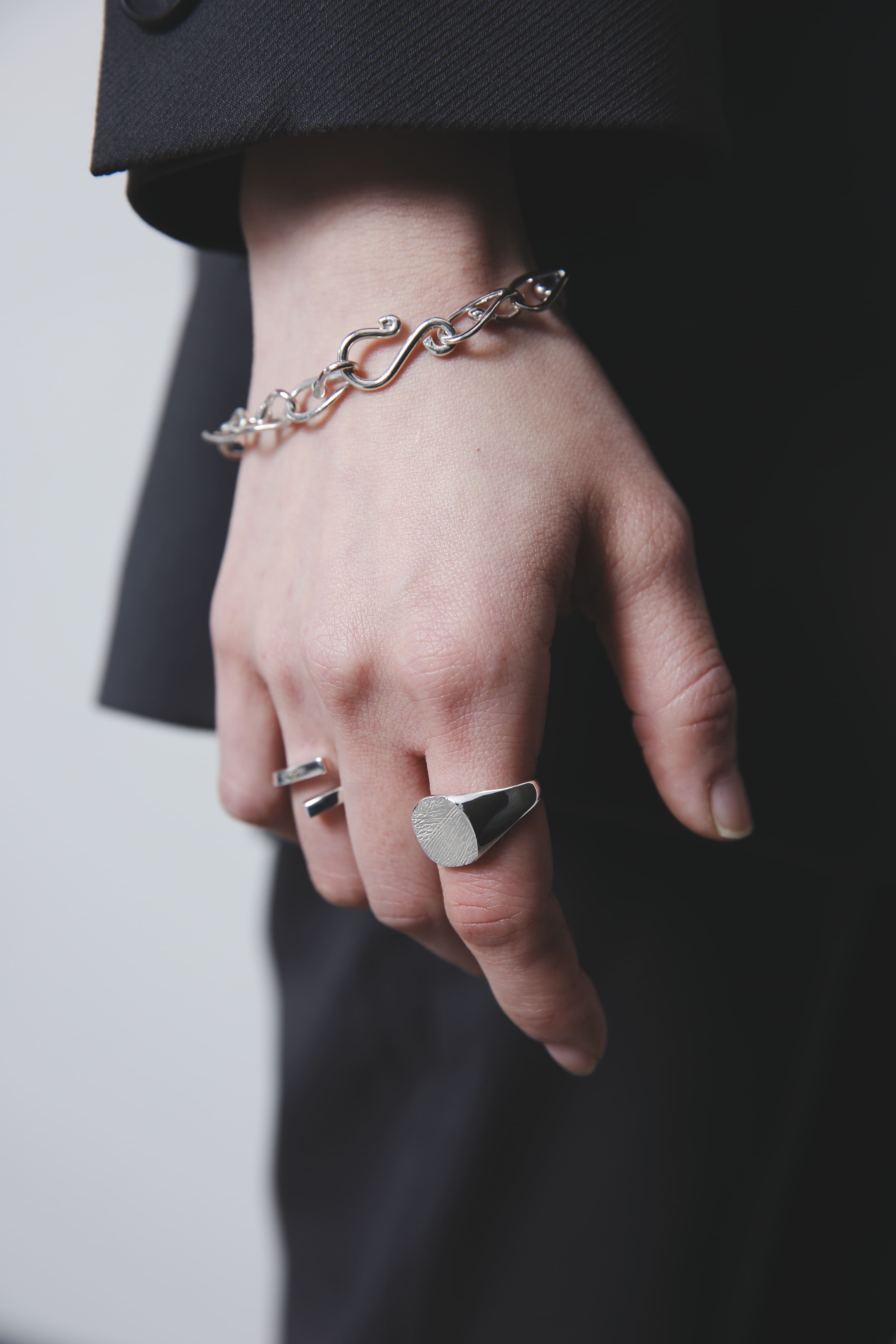 黒のジャケットを着用した手元。ねじれたような有機的なフォルムのシルバーチェーンブレスレットと、木目模様のシグネットリング、シンプルなオープンリングをコーディネート。無駄のない装いに控えめな輝きが映える。