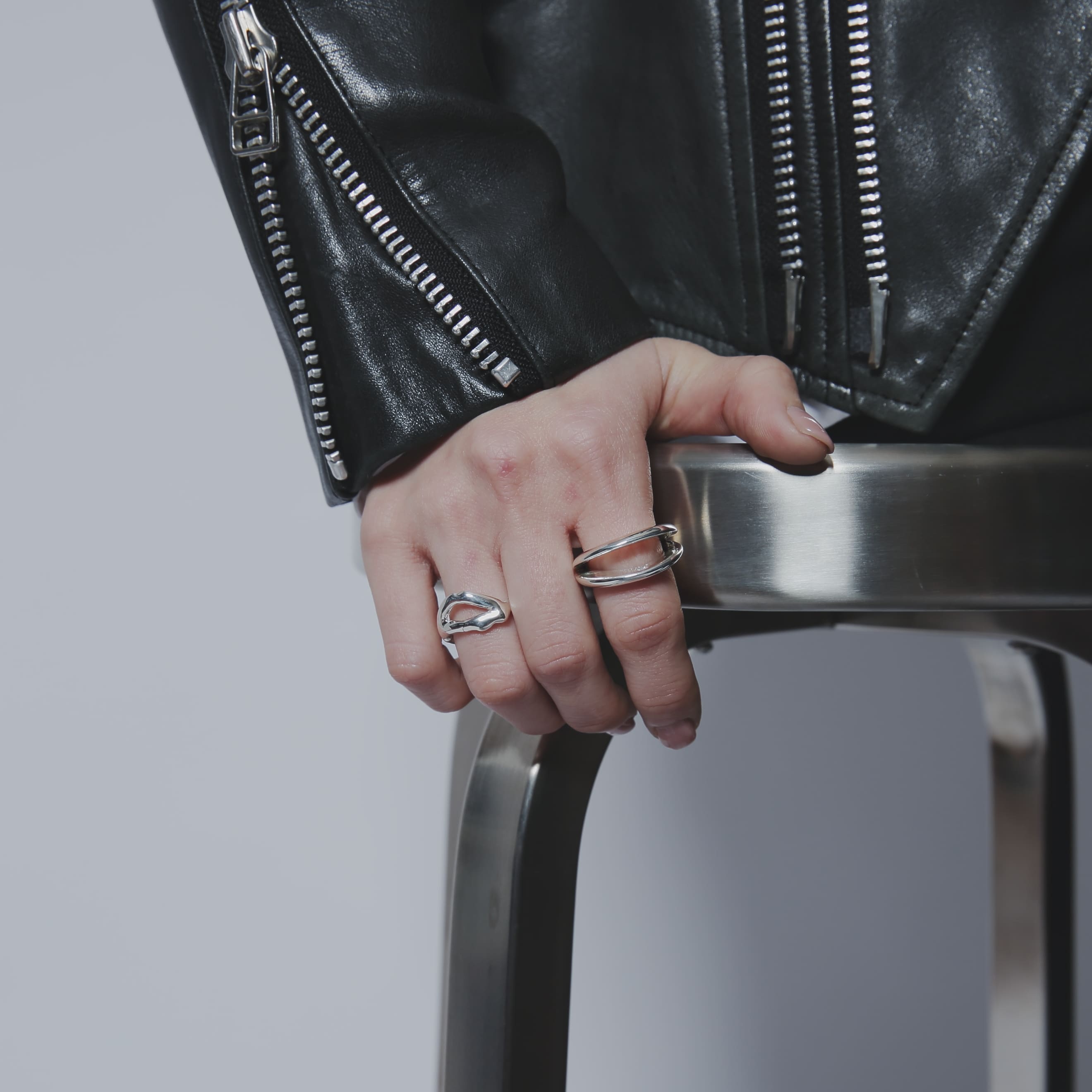 メタルのスツールに手を置いた、シルバー925リングの着用カット。ライダースジャケットのジップディテールと、シルバー925の有機的なフォルムが調和したモードな雰囲気。