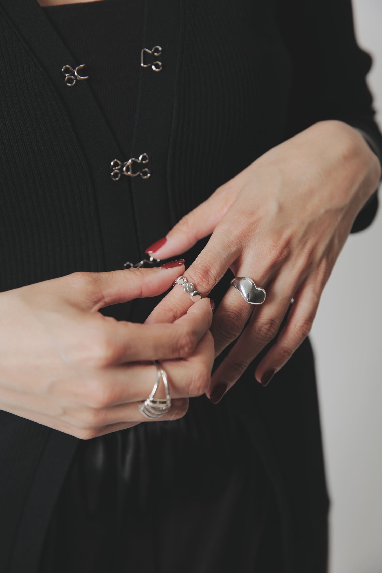 左手で右手のリングを整える動作を捉えた一枚。中指のシルバー925ハートリング、薬指のデザインリング、そして左手の親指付近に見える個性的なケージリングが、ジュエリーの重ね付けの楽しさを表現しています。