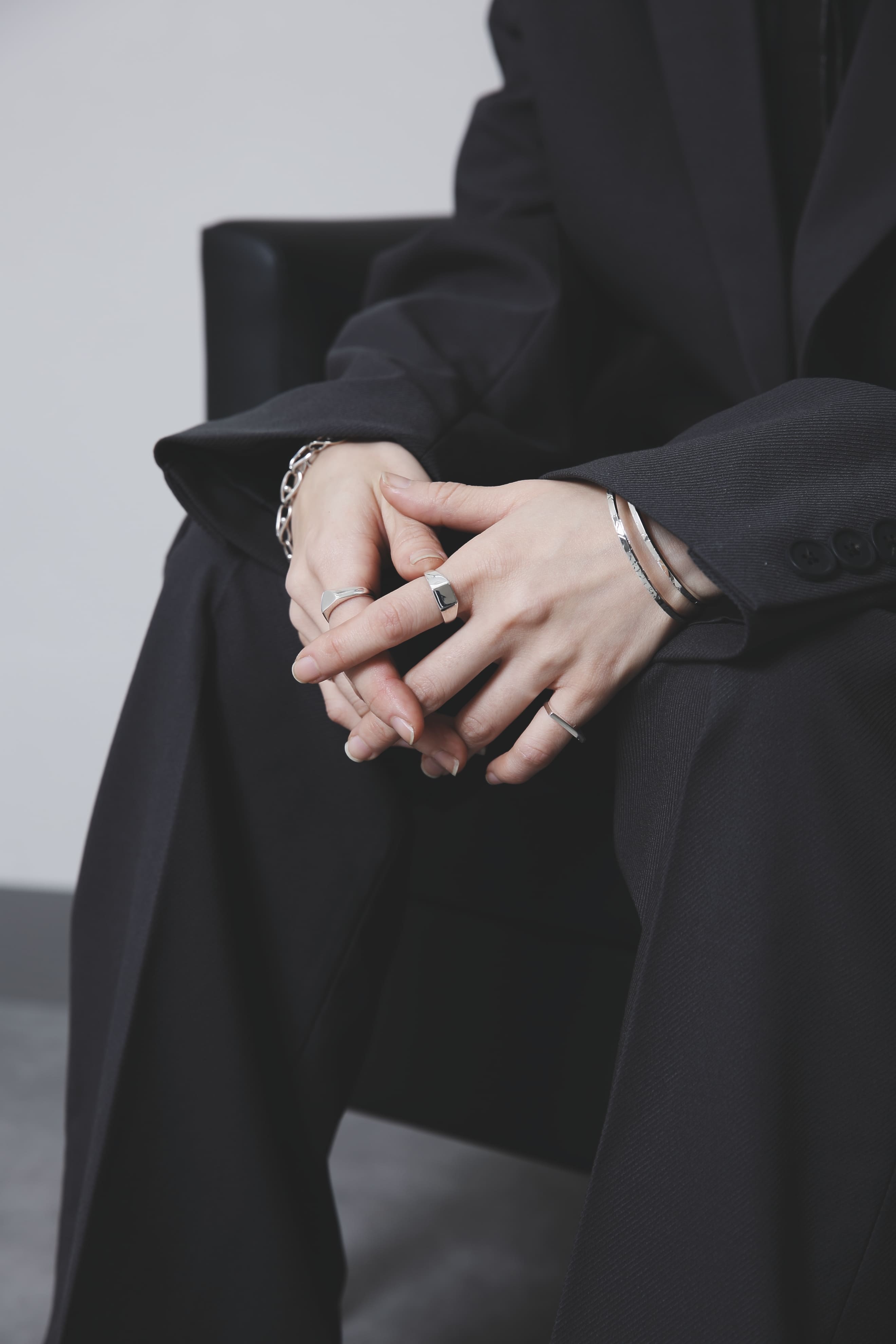 椅子に座り、指を組んだ手元のクローズアップ。スクエア型のシグネットリングが中心に輝き、シンプルな装いに知的な印象を添えている。