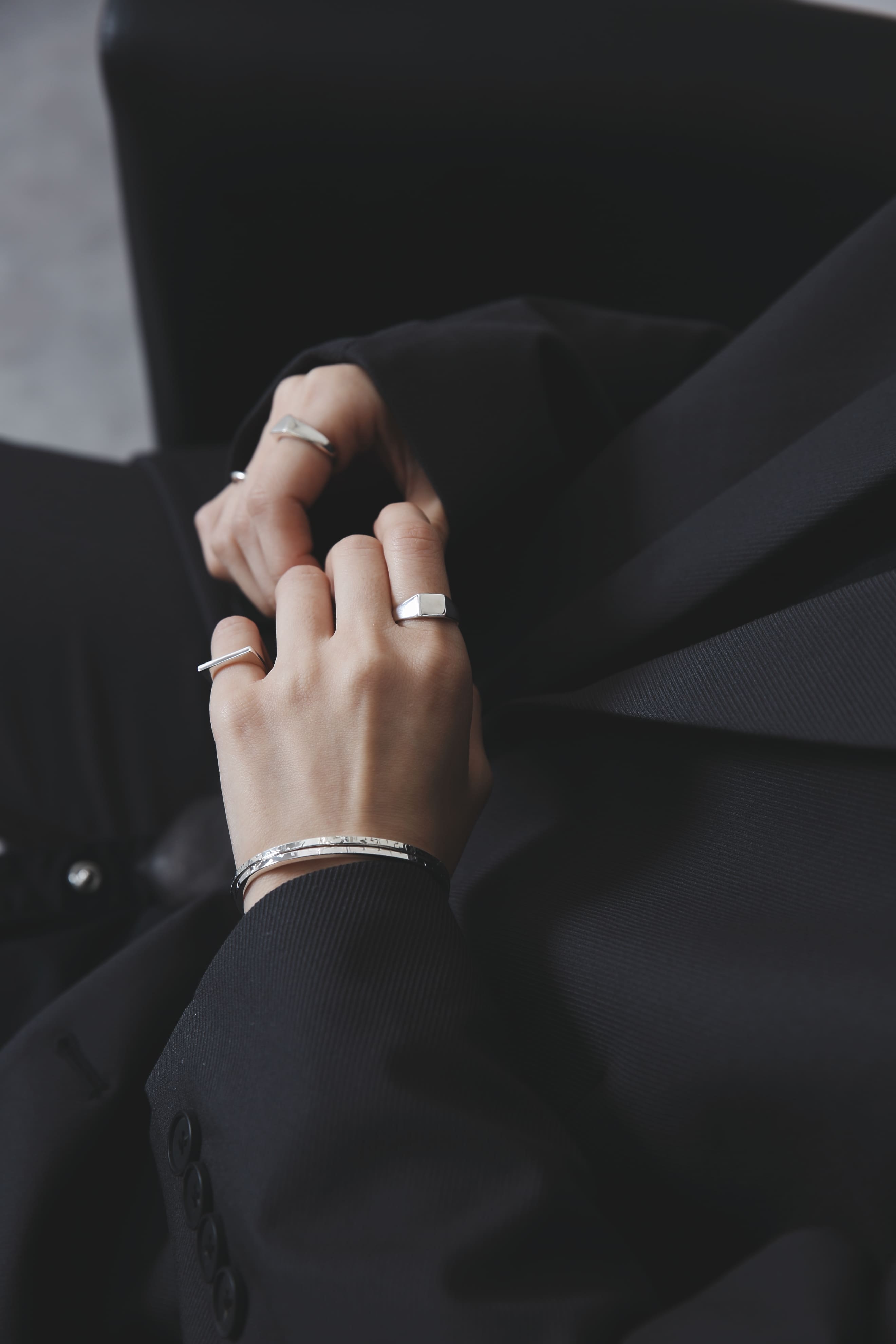 自然光のもと、黒いジャケットに合わせた手元のアップ。エッジの効いたスクエア型のシグネットリングが、装いの中で静かに存在感を放つ。