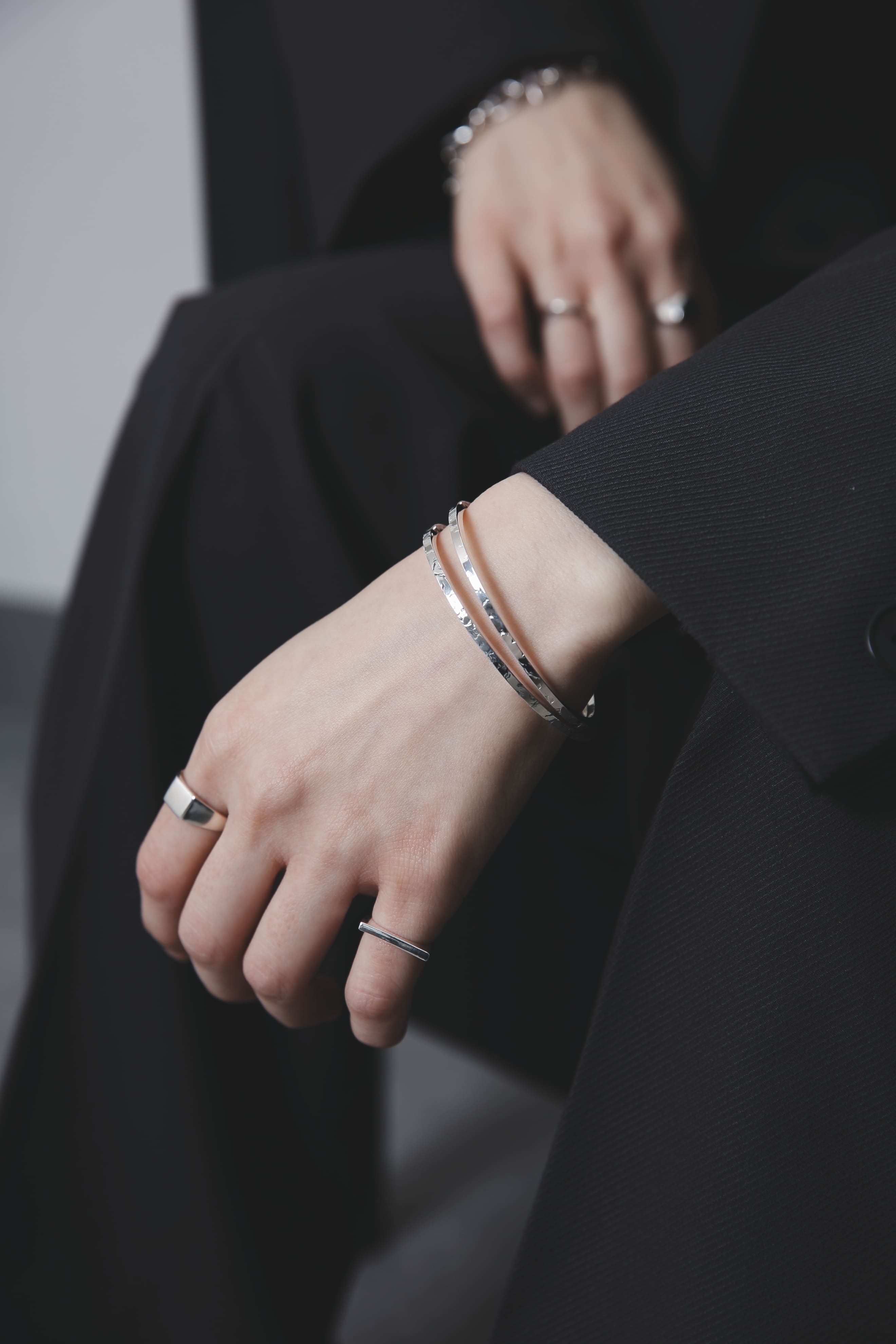 黒のセットアップに身を包んだ人物の手元に、シルバー925製の細身バングル2本と、モダンなデザインのリング2種を重ねづけしたスタイリングが映し出されている。シンプルながら洗練されたジュエリーの存在感が際立つカット。