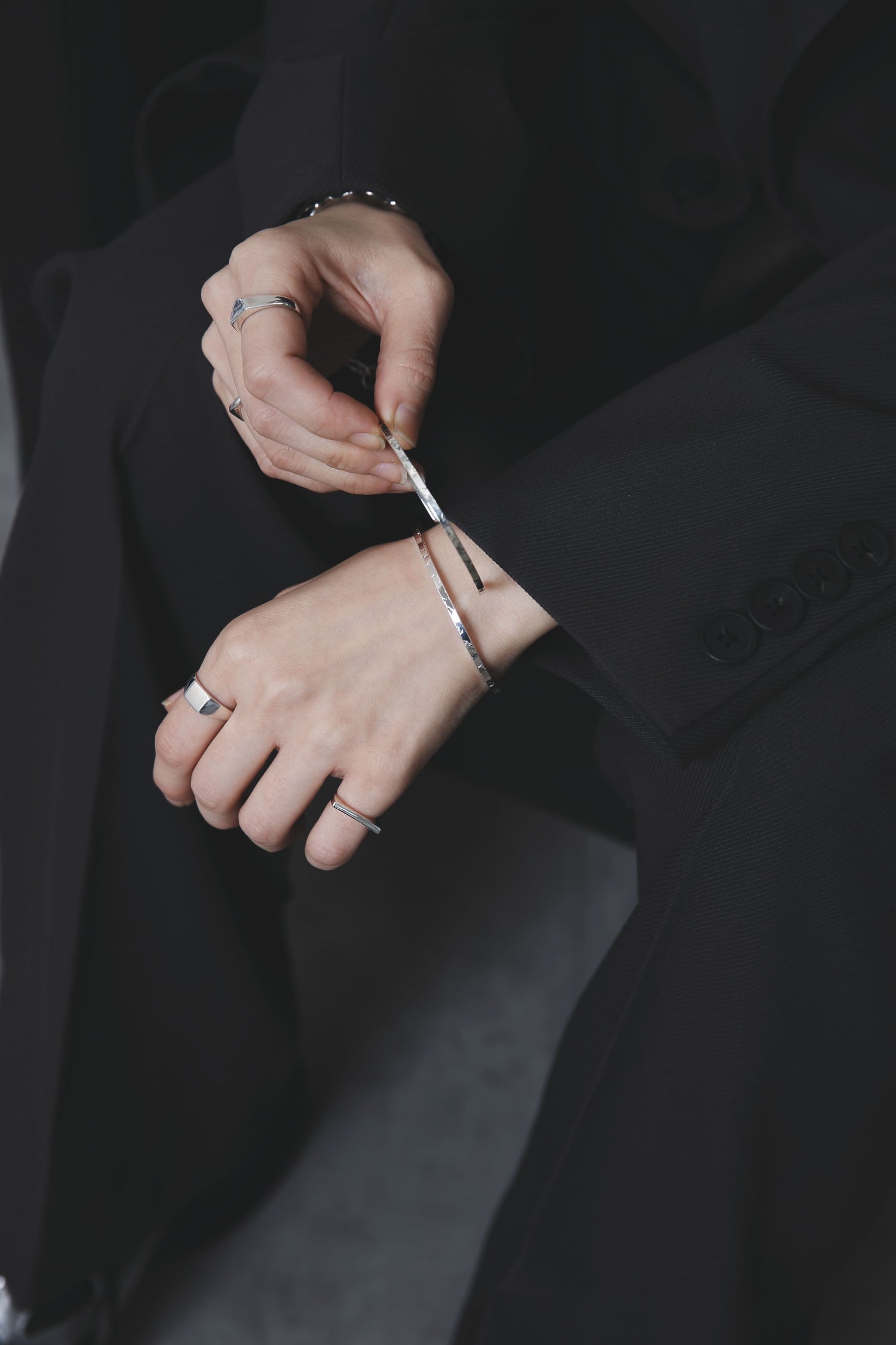 スーツ姿の人物が、手元に複数のシルバーアクセサリーを着けている様子。細身のバングルを2本と、存在感のあるリングを2本重ね付けし、洗練された雰囲気を演出している。