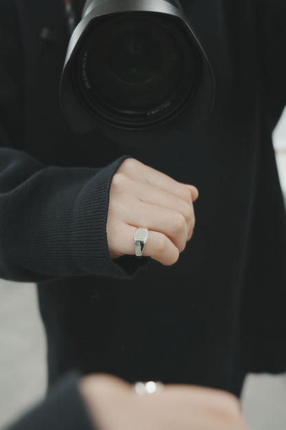 カメラを構える人物の手元を写したクローズアップ。鏡面仕上げのラウンドシグネットリングが、モノトーンの装いに上品な輝きを添えている